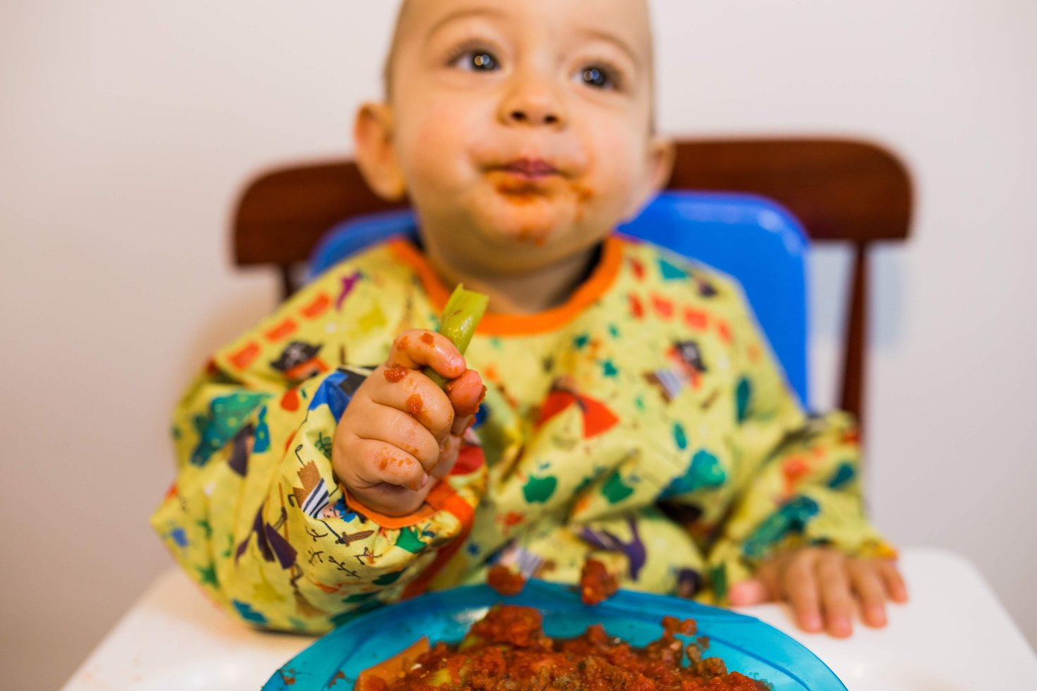 Bebê sentado na cadeira de alimentação vestindo babador BLW manga longa na cor amarela com desenhos das fábulas infantis. O bebê está comendo com as mãos um pedaço de vagem. O bebê com a boca suja de molho, mas a roupa protegida pelo babador BLW.