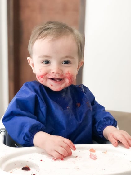 Bebê sentado na cadeira de alimentação com as mãos, boca e bochechas sujas de beterraba. A roupa está protegida por um babador BLW impermeável que conteve toda a sujeira da comida.