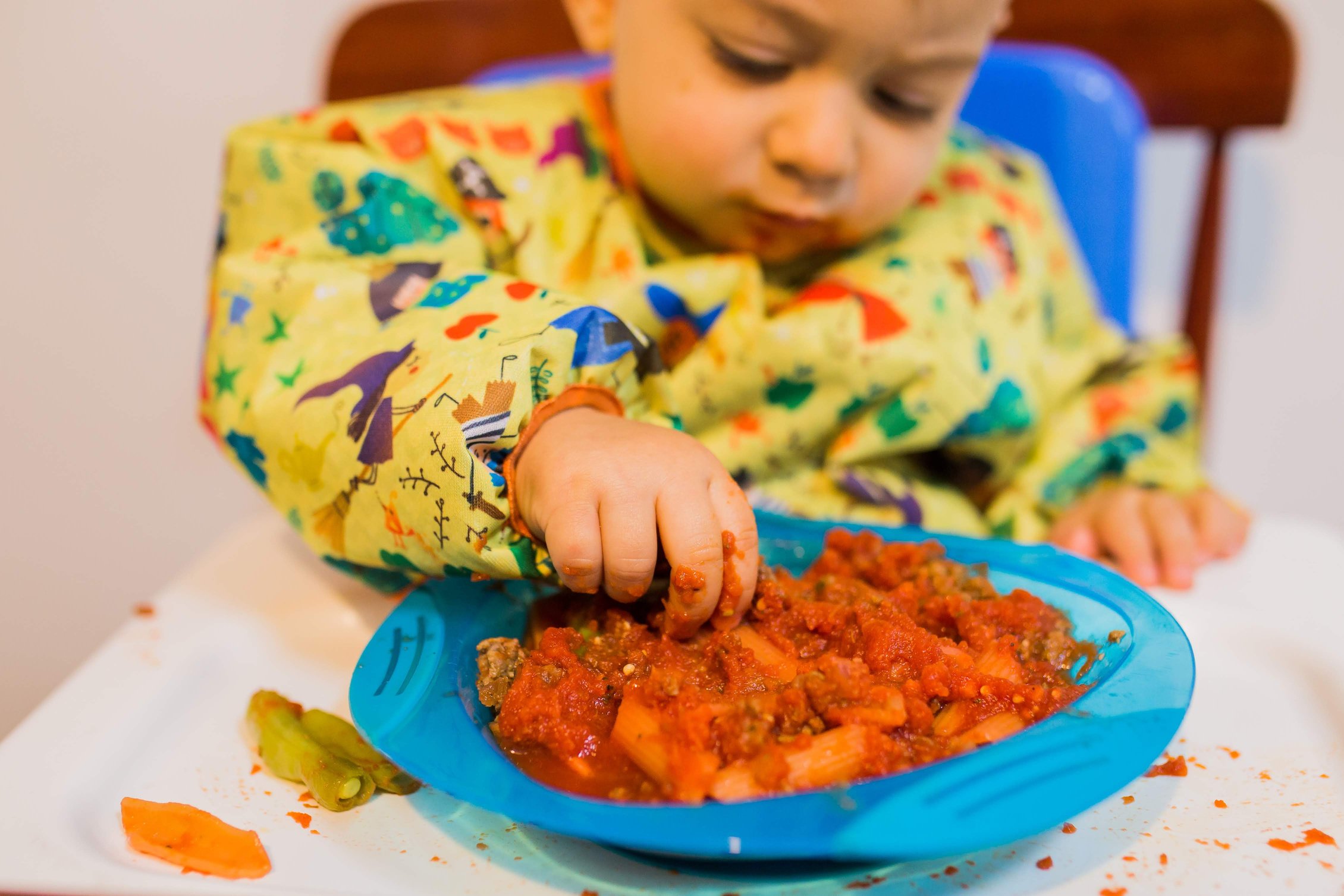 Apenas o corpo de um bebê sentado na cadeira de alimentação vestindo babador BLW manga longa na cor amarela com desenhos das fábulas infantis. O bebê está pegando com as mãos macarrão ao molho bolonhesa que está dentro de um pratinho azul.